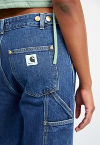 Primer plano de una persona que lleva pantalones vaqueros Carhartt azules, mostrando el bolsillo trasero con el logo y parte del brazo con un cordón de sudadera verde colgando.
