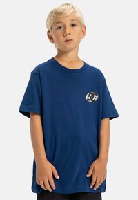 Blå bomullströja med rund hals, korta ärmar och en cirkulär grafisk logga på vänster bröst. Avslappnad passform, mjuk textur.