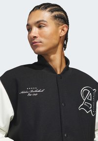 Black and white varsity jacket with embroidered "Adidas Basketball" logo and year "Est 1949." Features a high collar and snap buttons.