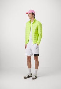 Chaqueta acolchada verde neón, camiseta blanca, pantalones cortos blancos con detalles en negro, zapatos grises y blancos con detalles neón, gorra rosa.