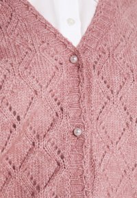 Detailní záběr na růžový pletený cardigan s diamantovými vzory a perlovými knoflíky přes bílou košili.
