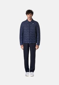 Giacca imbottita blu navy con colletto, caratterizzata da una chiusura con zip e un accento con marchio. Indossata sopra pantaloni scuri e sneakers bianche.
