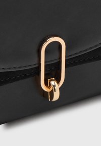 Černá kožená taška s uzávěrem zlatého oválného kroužku a háčkovým detailem, zobrazená zblízka na prošitém okraji klopy.