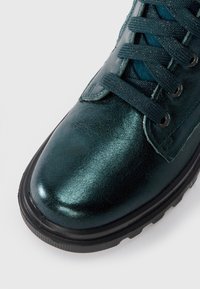 Bottine métallique verte avec une tige texturée, bout rond, semelle en caoutchouc noire et design à lacets avec des lacets sombres et des œillets.