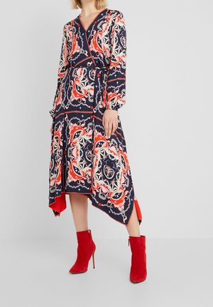 Femme portant une robe bleu marine à manches longues avec des motifs floraux rouges, blancs et beiges et un ourlet asymétrique, associée à des bottines rouges.