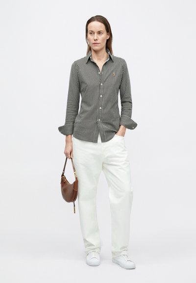 Fekete-fehér csíkos galléros ing, fehér, széles szárú nadrággal és fehér sportcipővel párosítva; barna válltáskát tart.