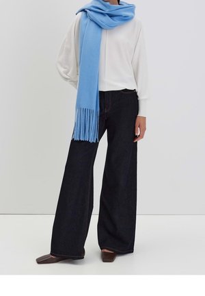 Femme portant un haut blanc à manches longues, un jean large foncé, des chaussures marron à bout carré et une longue écharpe bleu clair avec des franges.