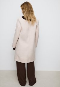Manteau en laine rose clair avec une coupe ajustée, des manches longues, des poches latérales et un col sombre, associé à un pantalon large marron.