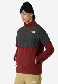 Joven con un gorro negro y una chaqueta de forro polar de dos tonos, roja y gris, con las manos en los bolsillos, de pie contra un fondo blanco.