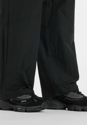 Zwarte sneakers met grijze accenten en het label "1000", gedragen bij losse zwarte broek op een neutrale achtergrond.