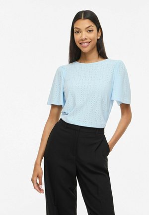 Femme portant un haut bleu clair à manches courtes perforé et un pantalon noir taille haute, souriante avec une main dans la poche.