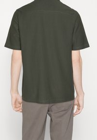 Uomo che indossa una polo a maniche corte verde scuro e pantaloni grigi, mostrato di spalle su uno sfondo bianco semplice.