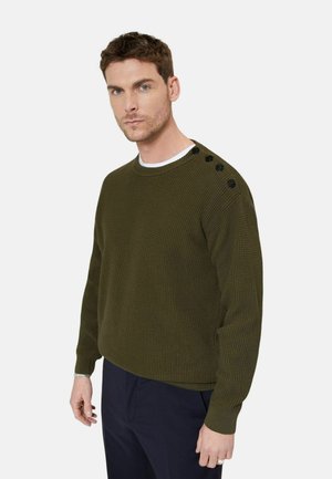 Mężczyzna ubrany w oliwkowo-zielony, teksturowany sweter dziany z czarnymi guzikami na lewym ramieniu, w połączeniu z ciemnymi spodniami, stojący na białym tle.