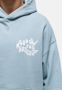 Sudadera con capucha azul claro hecha de tela suave que presenta un gráfico blanco audaz con el texto "GOOD FOR NOTHING" y detalles de mariposas impresos en el frente.