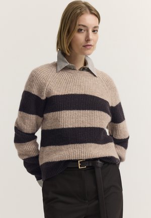 Junge Frau mit kurzem blondem Haar trägt einen beige-schwarz gestreiften Pullover über einem Hemd mit Kragen und schwarze Hose mit Gürtel.