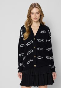 Zwarte cardigan met een wit "BIZUU" logo patroon, v-hals design, lange mouwen en gouden knopen. Draagt over een zwarte plissé rok.