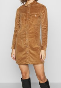 Brun kordflodskjolklänning med långa ärmar, krage och två bröstfickor. Har framknäppning och en figurnära silhuett.