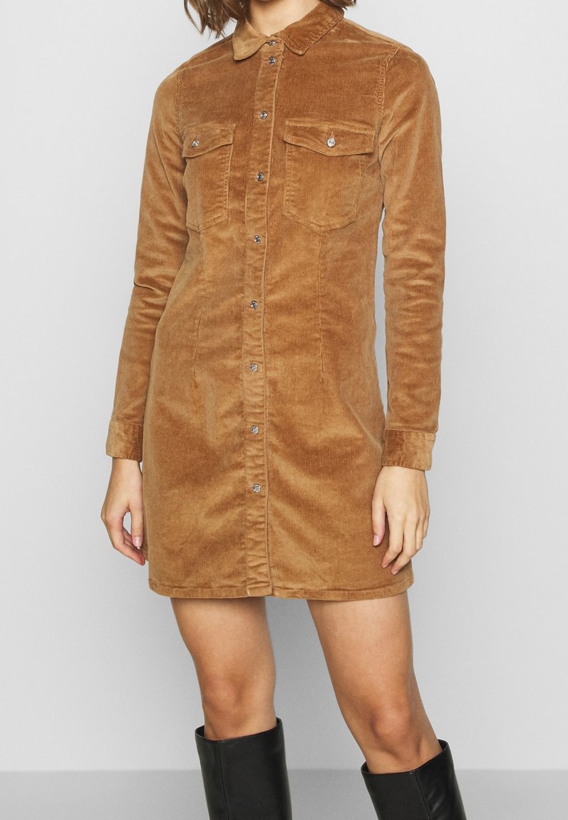 Brun kordflodskjolklänning med långa ärmar, krage och två bröstfickor. Har framknäppning och en figurnära silhuett.