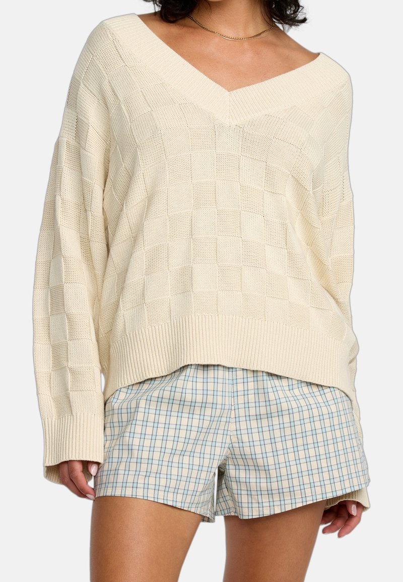Femme portant un pull en maille crème à col en V avec un motif à carreaux, assorti à un short à carreaux bleu clair et beige.