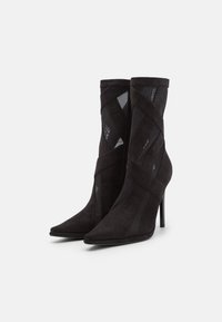 Bottines noires à talons aiguilles et bouts pointus, avec des panneaux en mesh et des lanières croisées en daim, sur fond blanc.