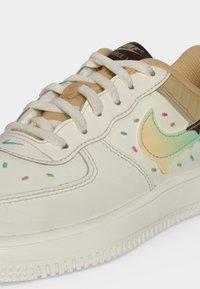 Άσπρο και μπεζ αθλητικό παπούτσι Nike με λευκά κορδόνια, πολύχρωμες κουφετένιες τελείες και λογότυπο Swoosh σε αποχρώσεις παστέλ στην πλευρά.