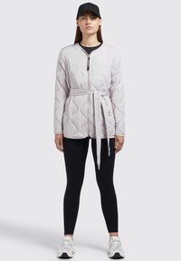 Lys grå quiltet jakke med bælte i taljen, rund halsudskæring og lynlås foran. Kombineret med sorte leggings og en sort kasket.