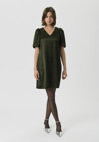 Rochie verde din satin, cu decolteu în V și mâneci bufante, până la genunchi. Asortată cu colanți negri și pantofi cu toc ascuțit argintii. Textură netedă.