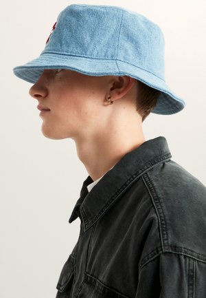 Sombrero tipo pescador de mezclilla azul con ala ancha y borde cosido. Lo lleva una persona que viste una camisa oscura.