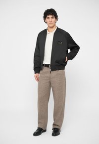 Μαύρο bomber jacket με χρυσό λογότυπο, φορεμένο πάνω από λευκό πόλο μπλουζάκι. Συνδυασμένο με ανοιχτό καφέ υφασμένα παντελόνια και μαύρα παπούτσια.