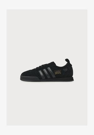 Baskets Adidas Samba noires avec des bandes noires, semelle texturée, lettrage doré et languette sur fond blanc uni.