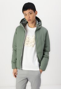 Grøn hættejakke med en glat overflade, med lynlås foran og en kontrastfarvet lys grå foring, båret over en hvid t-shirt.