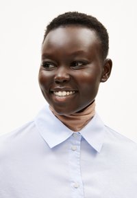 Lichtblauwe overhemd met kraag en een gladde textuur, voorzien van knopen, gecombineerd met een huidkleurige nekbedekking. Close-up van het gezicht, met een glimlach.