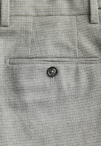 Hellgraue Hose mit kariertem Muster, ausgestattet mit einer Gesäßtasche und einem schwarzen Knopfdetail. Texturiertes Gewebe mit subtilen Webstrukturen.