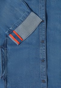 Manche d'une veste en denim bleu avec deux bandes rouges sur le poignet, boutons-pression noirs visibles et détail de couture de poche sur le côté gauche.