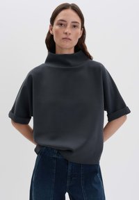 Donna con capelli scuri che indossa una maglietta nera a maniche corte e collo alto, in modo morbido, e jeans blu scuro, su uno sfondo chiaro e semplice.