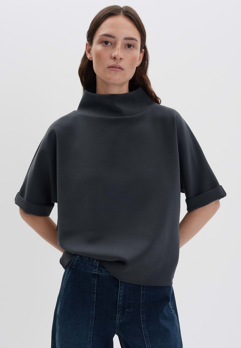 Donna con capelli scuri che indossa una maglietta nera a maniche corte e collo alto, in modo morbido, e jeans blu scuro, su uno sfondo chiaro e semplice.