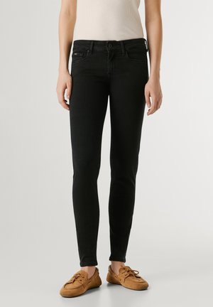 Person iført sorte skinny jeans, beige ærmeløs top og brune snørebånd i ruskindssko, stående mod en ensfarvet baggrund.