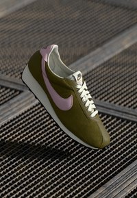 Sapatilha Nike com parte superior texturizada em verde oliva, swoosh rosa, atacadores brancos e sola branca. Colocada em frente a um fundo geométrico.