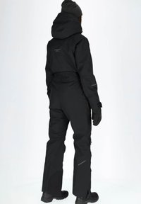 Zwarte waterdichte buitenkleding met een getailleerde pasvorm, capuchon, ritsen aan de zijkanten en getextureerde handschoenen. Verstevigde naden en stevige laarzen.