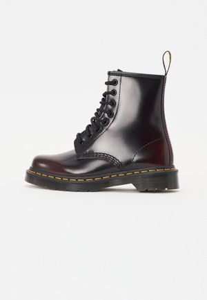 Bota de combate de cuero negro con cordones, costuras amarillas, suela gruesa y anilla trasera para tirar, mostrada en perfil lateral sobre fondo blanco.
