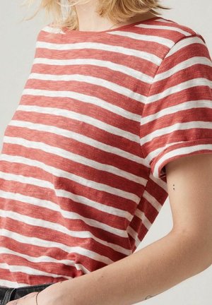 Persona que lleva una camiseta de manga corta con rayas horizontales rojas y blancas y un pequeño tatuaje de punto y coma en la parte superior del brazo.