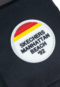 Tela negra con una insignia redonda blanca que presenta rayas rojas, amarillas y negras, y el texto "SKECHERS MANHATTAN BEACH '92."