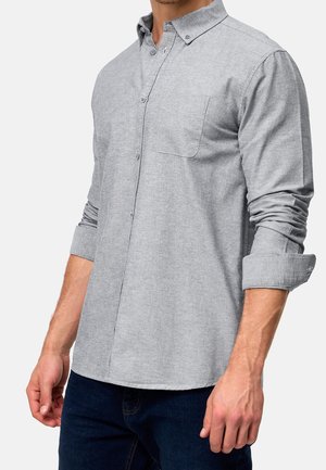 Chemise grise à manches longues et à boutons, avec une poche poitrine, un col structuré et des manches retroussées, fabriquée dans un tissu texturé.