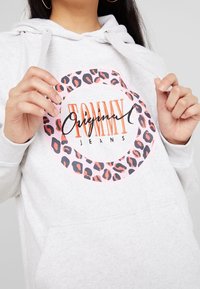 Žena v světle šedé mikině s kapucí s růžovým a černým leopardím potiskem v kruhu a oranžovým logem „Tommy Jeans Original“, ruce upravuj�í šňůrky mikiny.
