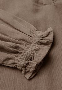 Primo piano di un tessuto marrone a coste con dettagli di polsini elasticizzati e arricciati su un indumento a maniche lunghe.