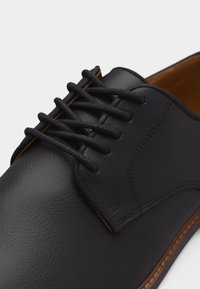 Μαύρο δερμάτινο παπούτσι φόρεμα με κορδόνια, στρογγυλερή μύτη και καφέ ραμμένη σόλα πάνω σε λευκό φόντο.