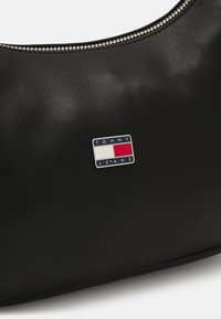 Bolso de mano de cuero sintético negro con forma redondeada, cierre de cremallera y un acento de logo plateado con detalles en rojo y blanco.