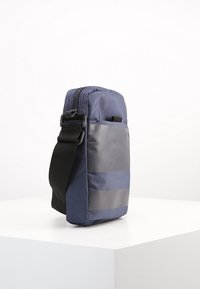 Borsa a tracolla blu in tessuto con chiusura a zip, dettagli grigi e tracolla regolabile. Forma rettangolare compatta con materiale testurizzato.