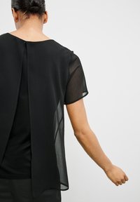 Černá blůza s průhledným zadním panelem a krátkými rukávy; má kulatý výstřih a mírný rozparek vzadu pro dodatečný detail.