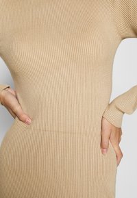 Gros plan d'une personne portant une robe en maille côtelée beige à manches longues, les mains posées sur les hanches, sur un fond uni.